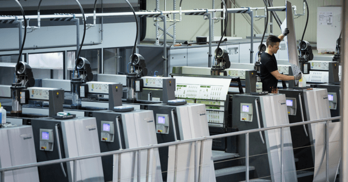 En arbetare driver en stor industriell tryckpress med flera kontrollpaneler och ark med tryckt papper som löper genom maskinen under en offsettryck-fabriksrundtur i en modern anläggning.
