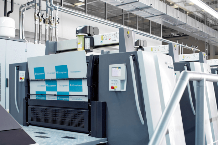 En modern industriell tryckpress i en väl upplyst anläggning som visar kontrollpaneler, bildskärmar och tryckplåtar med blå "Koenig & Bauer"-etiketter.