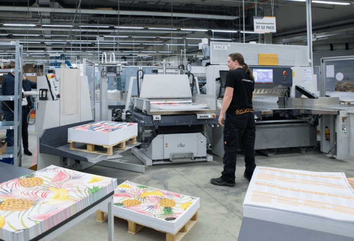 En arbetare driver en skärmaskin i en tryckeri, omgiven av stora tryckta ark med färgglada mönster. Högar med tryckt papper och industriell utrustning fyller den starkt upplysta arbetsytan.