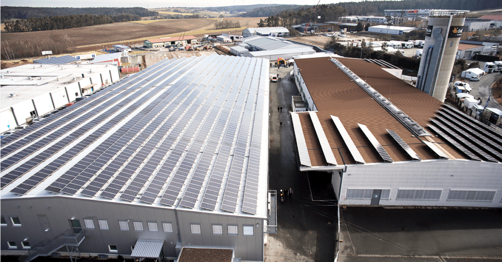 Flygfoto över ett industriområde med stora fabriksbyggnader; ett tak täckt med solpaneler, det andra med en brun metallyta. Omgivande fält, vägar och diverse uthus syns i bakgrunden.