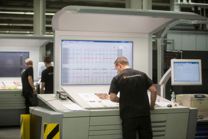 En man i en svart skjorta står vid en stor kontrollpanel med skärmar som visar grafer och diagram i en modern industriell tryckanläggning, medan andra arbetare syns i bakgrunden.