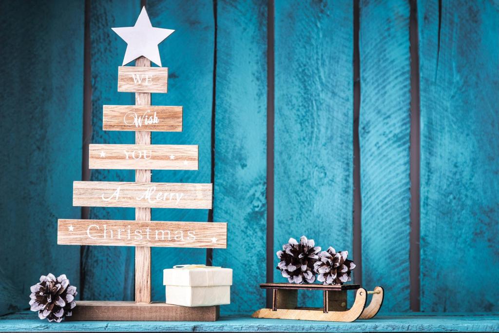 En julgran i trä med en stjärntopp och text, en liten vit presentask, tallkottar och en miniatyrsläde är arrangerade på en blå träyta med en blå träpanelbakgrund.