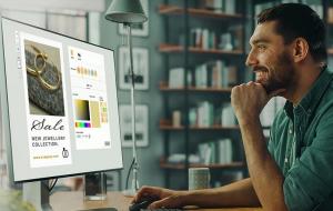 En leende man sitter vid ett skrivbord med hjälp av en dator och designar en onlineannons för en smyckeförsäljning. Datorskärmen visar smyckesbilder, färgalternativ och designverktyg. Hyllor och böcker syns i bakgrunden.
