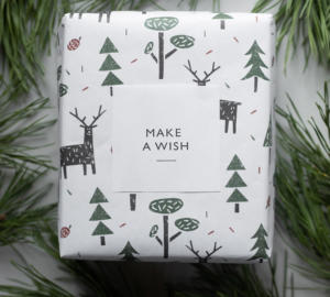 En gåva inslagen i vitt papper med gröna träd och hjortillustrationer, märkt med en fyrkantig tagg som läser MAKE A WISH, är omgiven av tallgrenar.