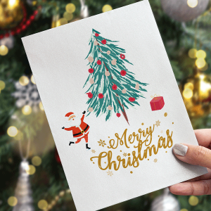 En hand håller ett julkort med illustrationer av ett dekorerat träd, jultomten, en present och guldtext som lyder "God jul", med en suddig julgran i bakgrunden.
